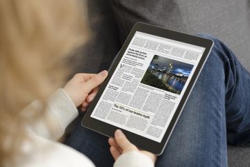 Over shoulder image of woman reading newspaper on tablet device.