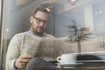 Bearded young business man seated with coffee reading a newspaper.