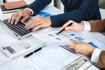 Two people reviewing report output and using their laptops. Notes and calculator on desk.