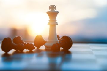 A white chess king on a chessboard, surrounded by fallen black pawns. The setting sun in back is casting a warm glow.