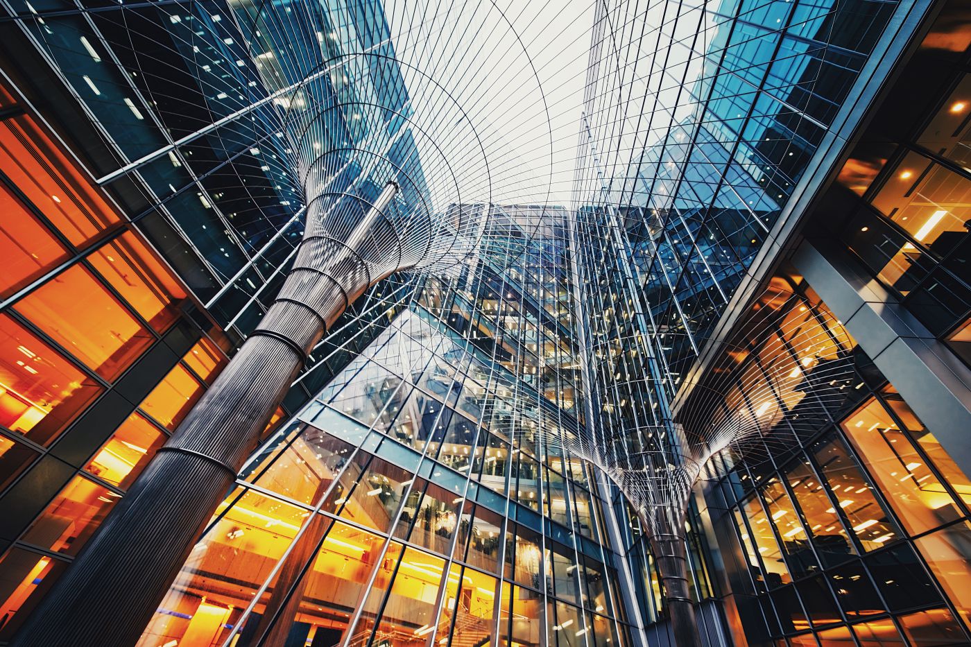 A modern, glass-fronted skyscraper with a unique metal sculpture in the foreground. The sky is visible through the glass panels.