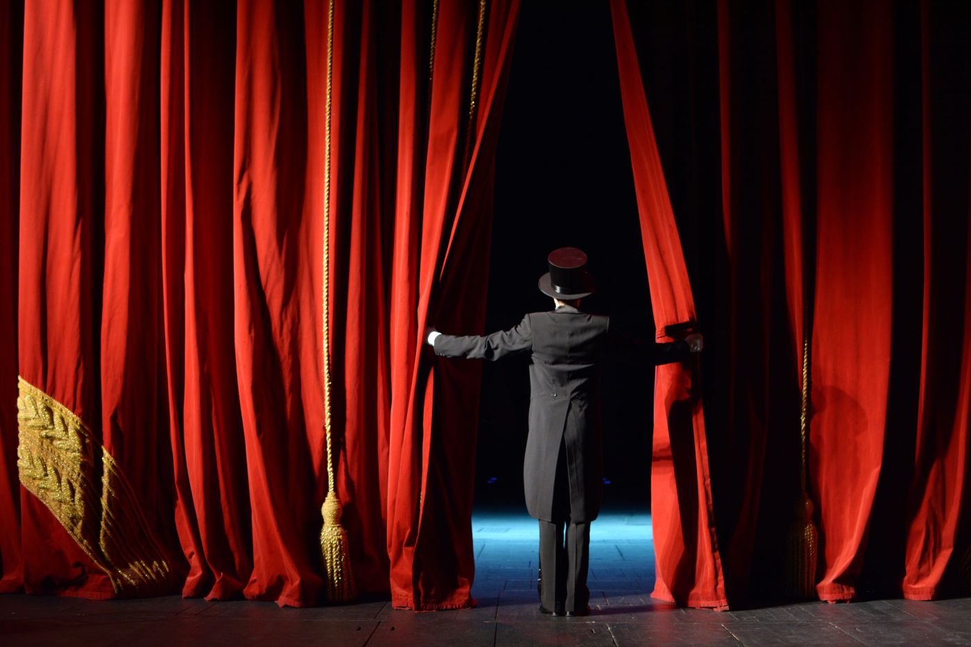 Woman in top hat and tails peeking through red stage curtains at audience.