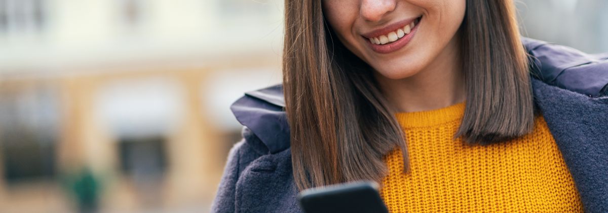 Smiling woman in a yellow sweater looking at her smartphone outdoors while wearing earbuds and a coat.