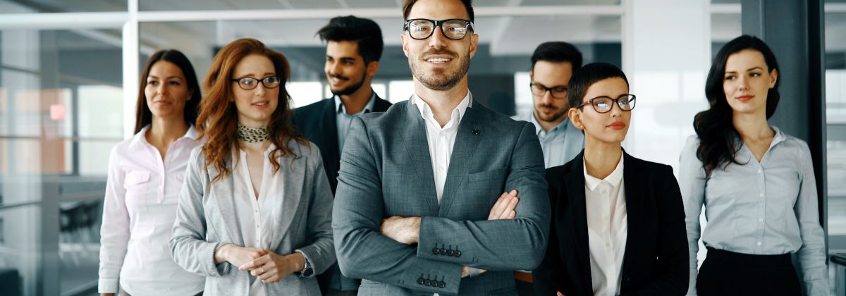 A group of smiling business professionals posing in an office, with one man in the front with crossed arms. 